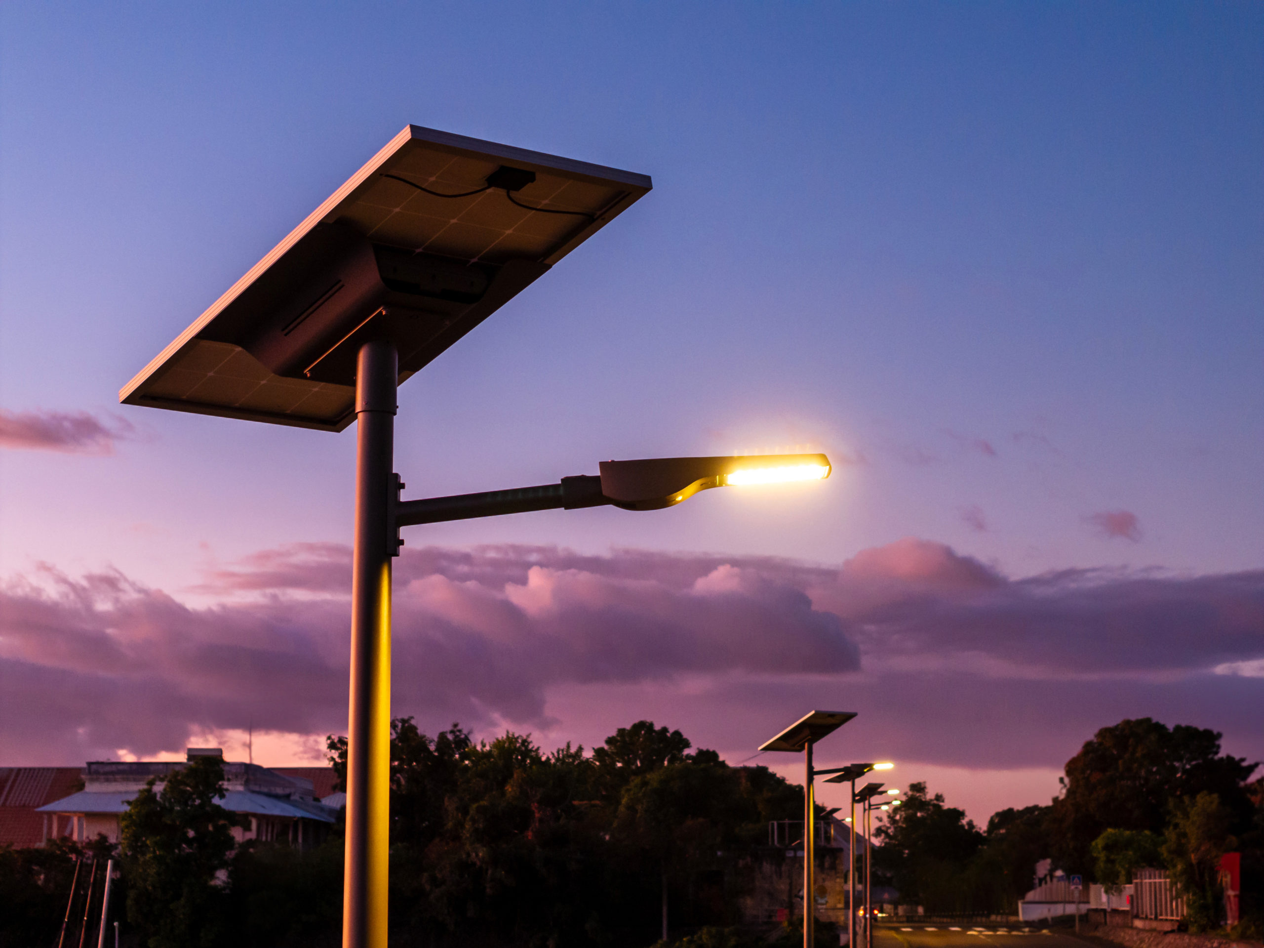 Luminaire public solaire allumé au crépuscule, équipé d’un panneau photovoltaïque en haut du mât, avec un ciel coloré en arrière-plan et des habitations entourées de végétation.